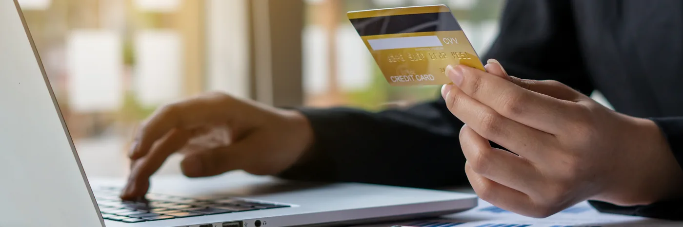 A person at a laptop holding a payment card in one hand, the other hand on the keyboard, partway through an online checkout.